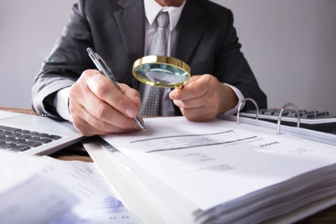 Homme d'affaires en costume-cravate analysant des documents financiers avec une loupe, stylo en main, assis à un bureau
